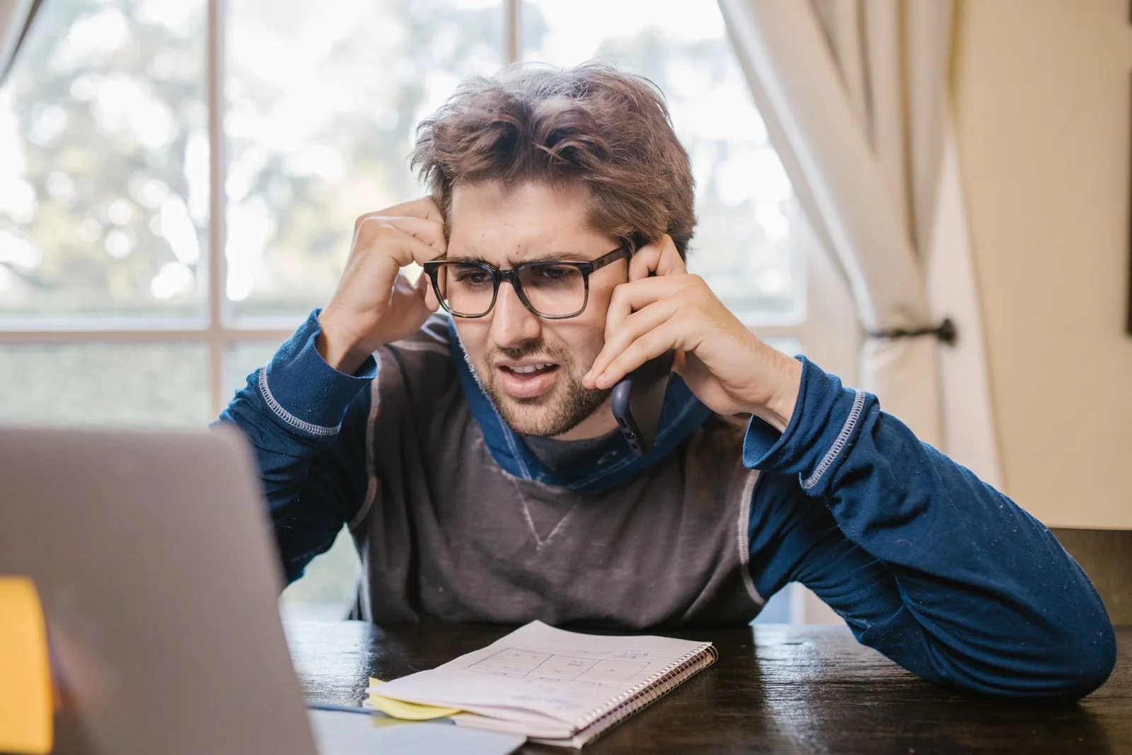 A man looking confused while using a laptop and smartphone together