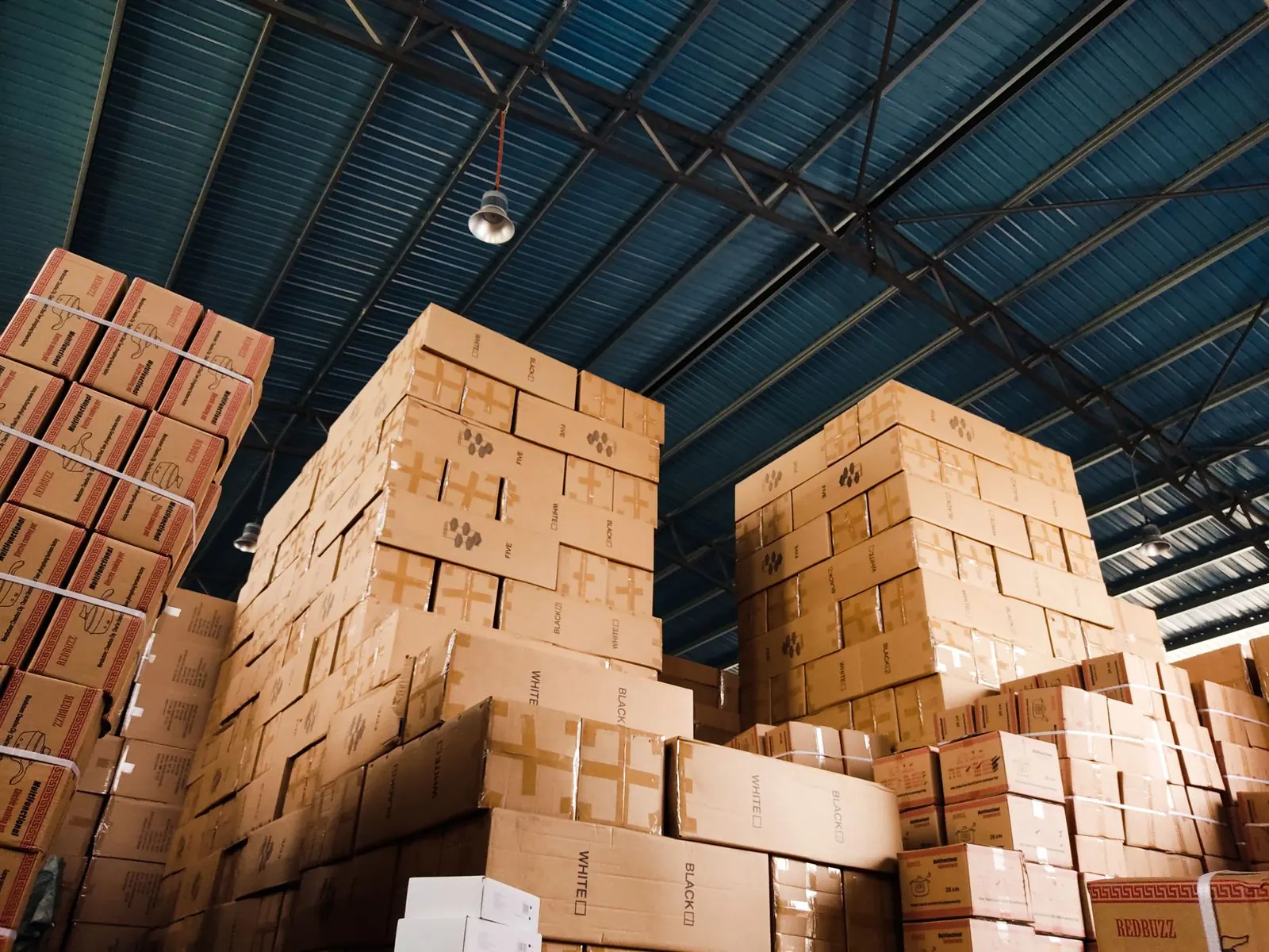 Tall stacks of cardboard boxes under the roof of a large warehouse