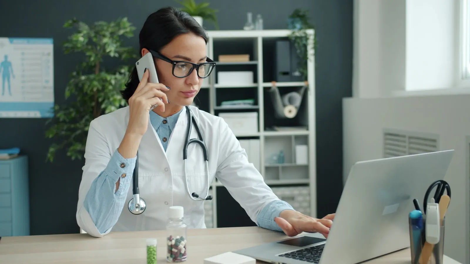 A doctor using a phone and a laptop at her desk in a medical office
