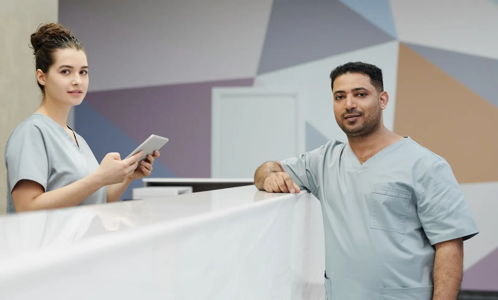 A receptionist and a colleague in scrubs at a clinic front desk